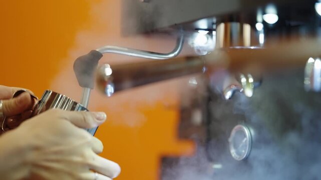 Close-up of a barista steaming milk with an espresso machine, frothing a stainless pitcher under a steam wand as vapor swirls against an orange background, creating microfoam