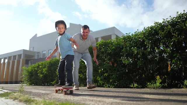 Boy learns skateboarding with father teaching near building outdoors. Child with helmet practices skating on path. Father helps boy learning skateboard. Dad teaches child skating safely.