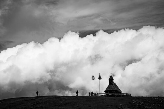 A dramatic black and white landscape featuring a small wooden chapel on a ridge, silhouetted against massive, towering white clouds with small figures of hikers nearby in Slovenia.