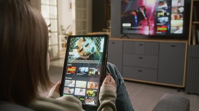 Over-shoulder shot of unrecognizable girl sitting on couch in living room, watching cyberpunk thriller trailer on streaming app on digital tablet, smart TV with movie selection menu in background