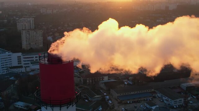 Smoke billows from industrial chimney at sunset light over cityscape.