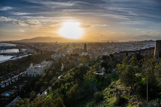 Aerial view of a hazy, golden sunset over Malaga city and harbor, seen from a distance with the historical Alcazaba fortress in the foreground.