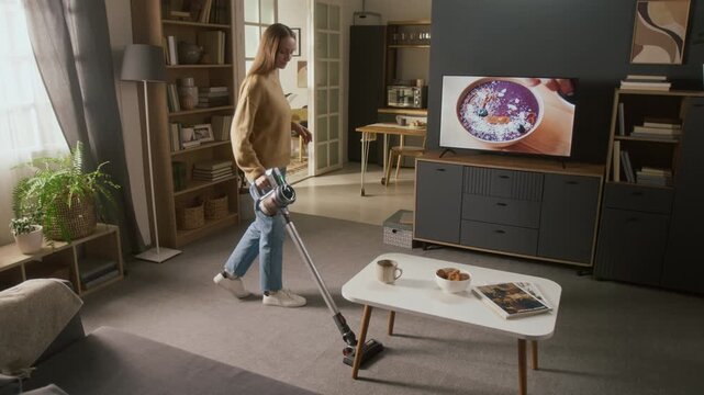 Wide arc shot of young Caucasian woman vacuuming floor in living room while watching cooking show on digital flat screen TV, female presenter decorating bowl of mousse and cutting ingredients