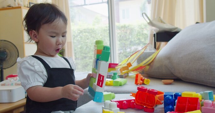 Engaged Asian toddler girl constructs a colorful tower using plastic toy blocks in a bright living room, focusing on early childhood development, creative playtime, and motor skills education.