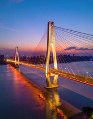 Obraz premium A stunning aerial view captures a modern suspension bridge illuminated at dusk, gracefully spanning a wide river with a vibrant cityscape visible in the background.