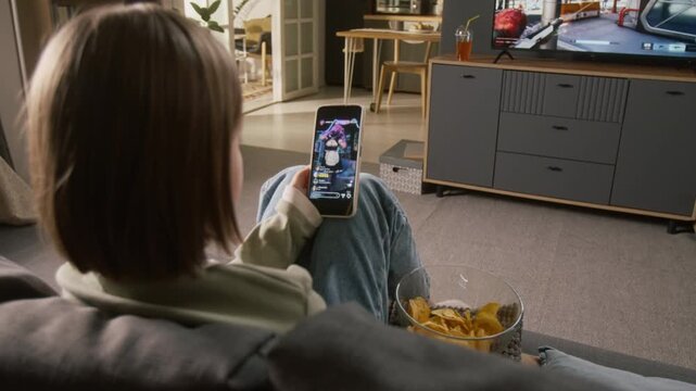 High-angle rear shot of young Caucasian girl sitting on couch at home, watching cosplay livestream on social media video platform, shooter gameplay on TV in background, childhood entertainment concept