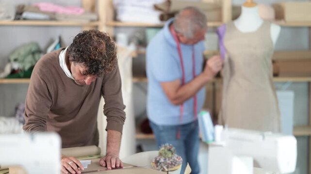 Adult man tailor drawing pattern on paper elderly man assistant sews on mannequin in workshop