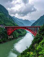 Obraz premium A striking red arch bridge gracefully spans a winding river, nestled amidst lush green mountains under a dramatic, cloudy sky.