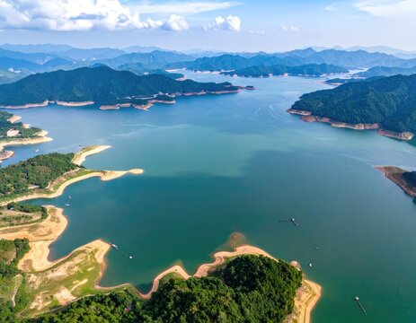 An aerial view showcases a vast reservoir nestled amidst rolling green hills and distant mountain ranges under a bright blue sky with scattered clouds.