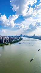 Obraz premium An aerial view showcases a sprawling cityscape nestled along a wide river, framed by a vibrant blue sky dotted with fluffy white clouds and a modern bridge connecting the urban landscape.