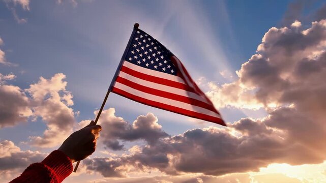 Hand holding american flag waving in the sky with clouds and sun rays