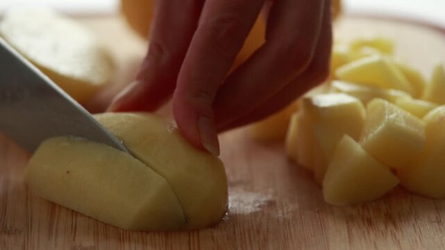 Close-up of woman's hands slicing and peeling fresh raw potatoes on a rustic wooden cutting board in bright natural light