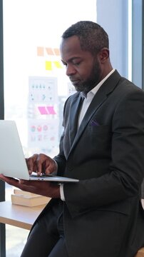 A focused African American businessman in a suit works diligently on his laptop while sitting near a window with charts on the wall