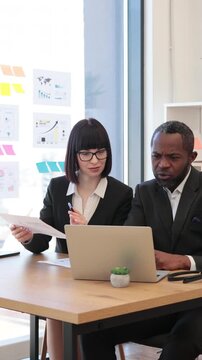 A woman and an African American man collaborate on a project using a laptop and documents in a modern office setting