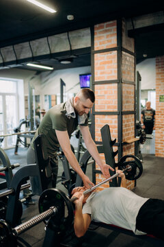 A man lies on a bench pressing weights while another man assists him in a gym. The atmosphere is busy with people working out and equipment around.