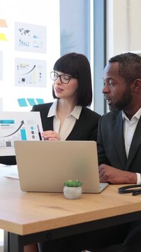 A woman in glasses presents a chart to an African American man during a business meeting with a laptop and charts on the wall