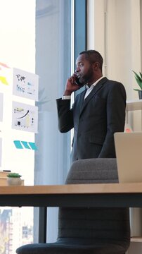 An African American businessman in a suit talks on his phone while looking at charts and graphs on a glass wall
