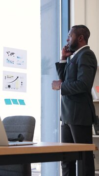 An African American businessman in a suit talks on his phone while pointing at charts and graphs on a glass wall in an office setting
