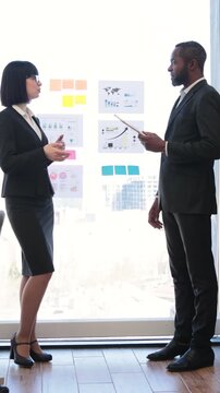 A woman and an African American man in business attire discuss charts and graphs on a glass board during a meeting