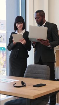 A woman and an African American man in business attire collaborate using a tablet and laptop in a modern office setting