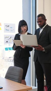 A woman and an African American man in suits collaborate on a project using a laptop and tablet in a modern office setting
