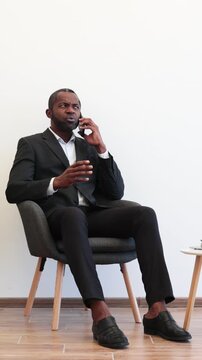 An African American man in a suit sits in a chair while talking on his phone with a perplexed expression, gesturing with his hand