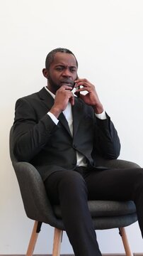 An African American man in a suit sits in a chair while talking on his cell phone, looking upwards thoughtfully