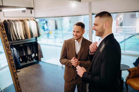 Two men stand in a clothing store looking at themselves in a mirror. They are trying on different suits and smiling. A rack of suits is visible in the background.