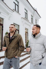 Two men walk on steps outside a white building in winter. They drink coffee from cups while wearing jackets. Snow is on the ground around them as they talk.
