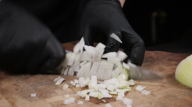 A close-up, high-angle shot of a chef wearing black nitrile gloves expertly dicing a fresh white onion on a rustic wooden cutting board. The video captures the precision and technique used in professi