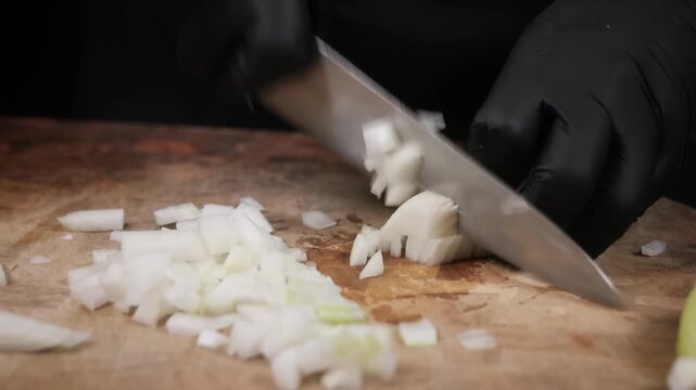 A close-up, high-angle shot of a chef wearing black nitrile gloves expertly dicing a fresh white onion on a rustic wooden cutting board. The video captures the precision and technique used in professi