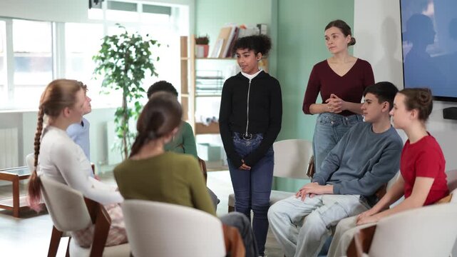 Teen group sit in circle and listen to African American girl classmate speaking, teacher watching. Acting masters courses, stage speech courses, public speaking courses