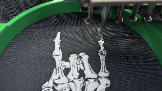 Industrial Multi-Needle Embroidery Machine Stitching White Skeleton Hand Rock Sign Logo on Black Fabric Macro Close Up Handheld Slow Motion Authentic High Angle Production Manufacturing View
