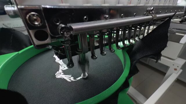 Industrial Multi-Needle Embroidery Machine Head Rapidly Stitching White Design on Black Fabric in Professional Garment Factory Workshop Macro Close Up Handheld Slow Motion Authentic Textile Scene