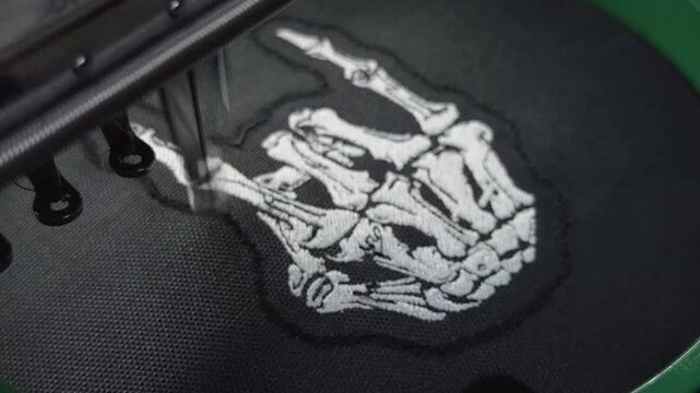 Macro Close Up Industrial Computerized Embroidery Machine Stitching Final White Thread Border on Skeleton Hand Rock Sign Design Black Fabric Handheld Slow Motion Authentic Production