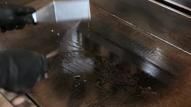 High-angle, close-up shot of a dirty commercial stainless steel flat-top grill being scraped and cleaned. The footage captures the satisfying process of removing grease and burnt food residue from the