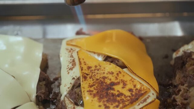 A close-up shot of a chef using a kitchen torch to melt a slice of orange cheddar or American cheese over seasoned, shredded beef on a flat-top grill. The meat is being prepared for making traditional