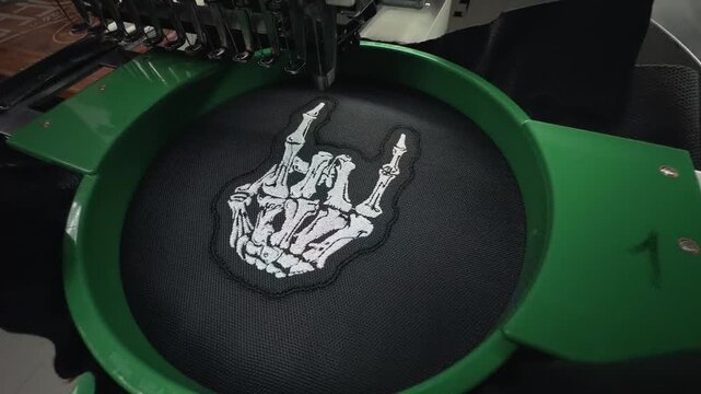 Wide Angle High View Industrial Computerized Embroidery Machine Stitching White Skeleton Hand Rock Sign Logo on Black Fabric Handheld Slow Motion Authentic Production Scene
