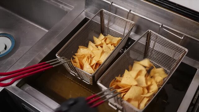 A close-up shot of a commercial deep fryer shows a batch of corn tortilla chips being lifted from bubbling hot oil. Another basket full of crispy, golden chips sits next to it.