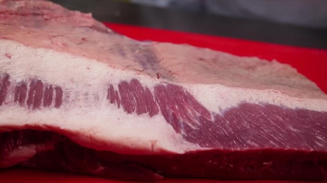 A close-up shot of a person using a sharp knife to carefully trim the thick fat cap from a large, raw beef brisket on a red cutting board. This preparation step is essential for proper cooking, such a