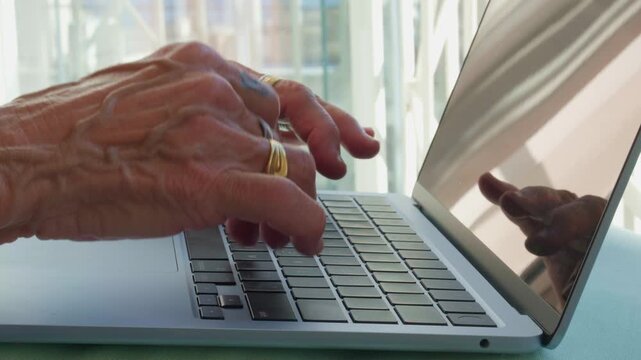 Close up of senior hands with rings typing on a laptop keyboard. Concept of elderly digital literacy, remote work for seniors, silver economy, and modern technology use in a home office