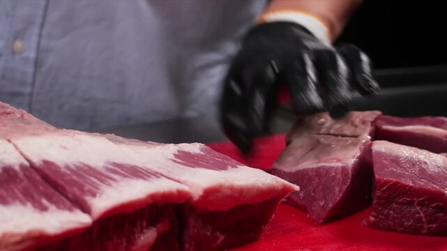 A detailed close-up shot of a person wearing black gloves using a sharp chef's knife to expertly slice a large, fresh, raw red beef steak on a vibrant red plastic cutting board.