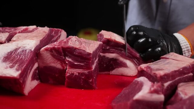 A close-up shot captures a professional butcher's hand, covered in a black safety glove, using a sharp knife to precisely slice a large slab of raw, marbled beef short ribs on a bright red cutting boa