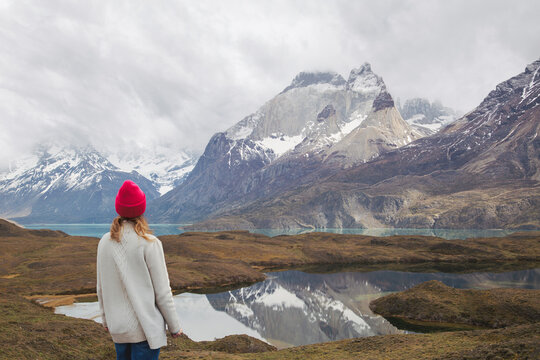A person wearing a bright red beanie stands before a serene glacial lake, watching the dramatic reflection of the snow-dusted Cuernos del Paine mountains in Patagonia.