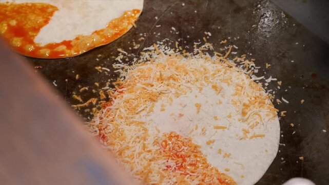 A close-up shot of a chef's hand in a black glove using a spoon to spread vibrant red birria sauce or consomm&eacute; onto round white tortillas on a hot commercial griddle.