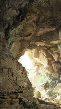 A person walks through a rocky cave with sunlight illuminating the stones. Plants grow inside, adding a touch of green to the dry rocky walls. Shadows create a mysterious atmosphere.