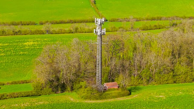 Rural cell tower with multiple antennas rising above trees and green fields with small utility building. Mobile network base station with countryside coverage and wireless signal distribution. Telecom