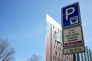 Obraz premium Paid parking zone sign in the foreground against a modern triangular flatiron-style office building and clear blue sky. Urban infrastructure and city traffic regulation concept.