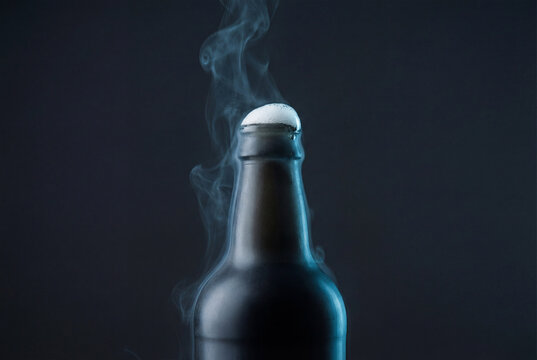 Cold beer bottle with foam and mist on dark background