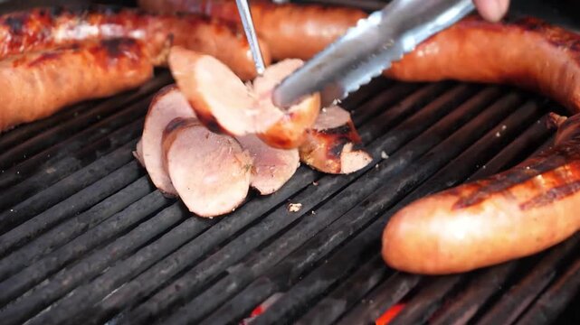 close-up shot of juicy, golden-brown sausages (kielbasa or bratwurst) grilling on a hot, cast-iron barbecue grill over charcoal. A hand uses metal tongs to adjust the sausages, one of which has cut ma